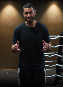 Man wearing a black t shirt speaks and gestures in a gym beside a boxing ring